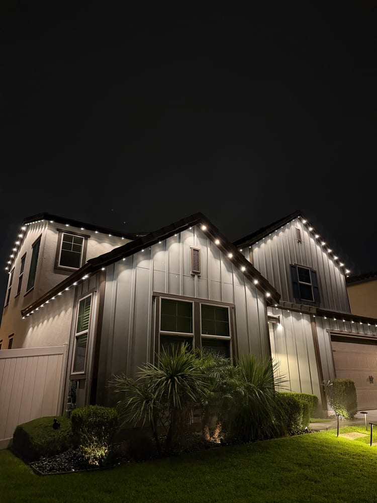 Board-and-batten home at night with bright permanent eave lights on the gables, landscape path lights and lit palms
