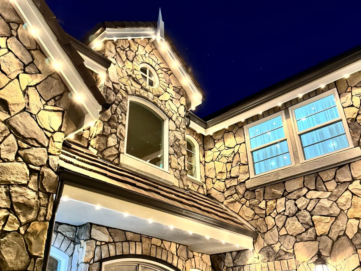 Stone home at dusk with warm white eave lights on its gables and a wreath on the front door