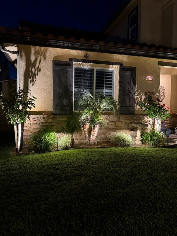 Front of a home at night with uplit shrubs, a small palm and landscape lights along the lawn