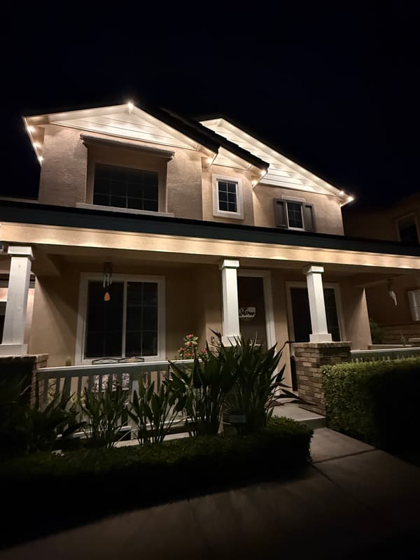 Two-story home with a columned front porch at night, warm white lights along the gables and porch roof